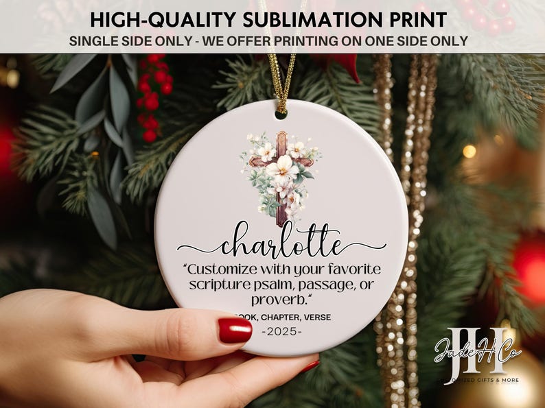 May include: A round, white ceramic ornament with a floral cross design and the name "Charlotte" in script. The ornament includes the text "Customize with your favorite scripture psalm, passage, or proverb."