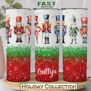May include: Three stainless steel tumblers with a red and green glitter design. The tumblers feature a watercolor illustration of nutcracker soldiers. The middle tumbler has the name "Cailtyn" written on it. The tumblers are part of a holiday collection.