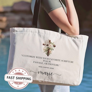 May include: A cream-colored canvas tote bag featuring a graphic of a wooden cross adorned with white roses and green leaves. The text reads: "CUSTOMIZE WITH YOUR FAVORITE SCRIPTURE PSALM, PASSAGE, OR PROVERB." followed by "BOOK, CHAPTER, VERSE" and the name "marie".