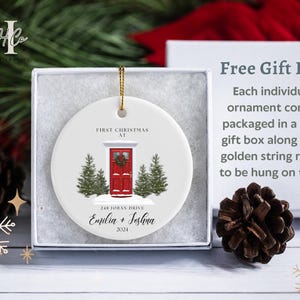 May include: White ceramic Christmas ornament with a red door, green trees, and the text "First Christmas at 248 Joban Drive Emilia + Jeshua 2024".