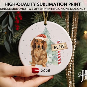 May include: A round Christmas ornament featuring a dachshund wearing a Santa hat, with a sign that reads "ELFIE" and the year "2025". The ornament has a gold string for hanging and is decorated with a Christmas tree.