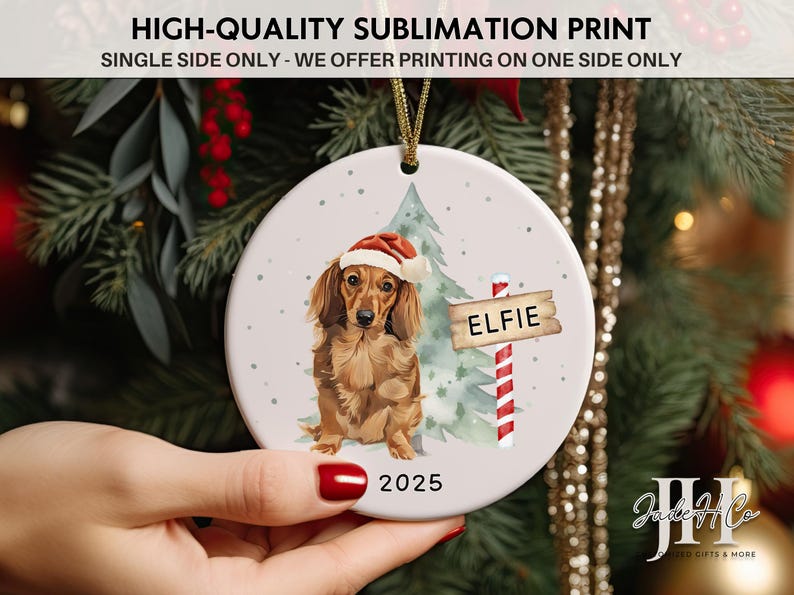 May include: A round Christmas ornament featuring a dachshund wearing a Santa hat, with a sign that reads "ELFIE" and the year "2025". The ornament has a gold string for hanging and is decorated with a Christmas tree.