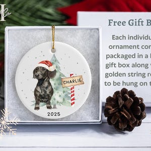 May include: A round Christmas ornament featuring a dachshund wearing a Santa hat, standing next to a Christmas tree and a sign that reads "Charlie". The ornament is packaged in a gift box with a golden string for hanging. The year 2025 is printed on the ornament.