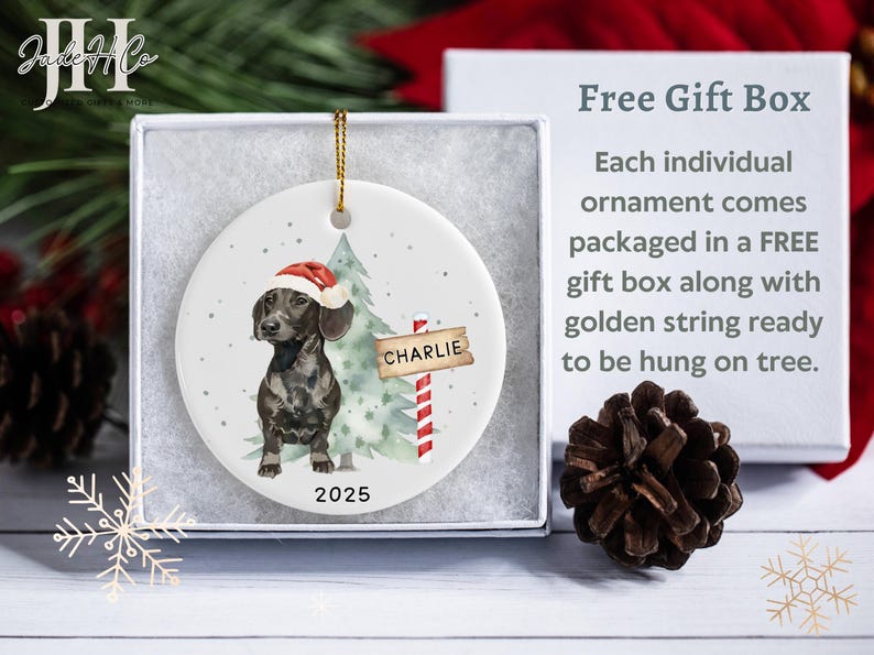May include: A round Christmas ornament featuring a dachshund wearing a Santa hat, standing next to a Christmas tree and a sign that reads "Charlie". The ornament is packaged in a gift box with a golden string for hanging. The year 2025 is printed on the ornament.
