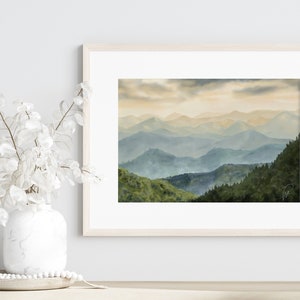 以下が含まれることがあります： 青と緑のぼやけた色合いの山脈を描いた、額縁に入った水彩画。山々は重なり合っており、手前の山はより詳細に描かれ、奥の山は背景に溶け込んでいく。