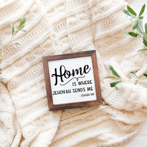 May include: A square wooden sign with the text "Home is where Jehovah sends me - Isaiah 68" in black script. The sign is set against a cream-colored knitted blanket, with sprigs of greenery on either side.