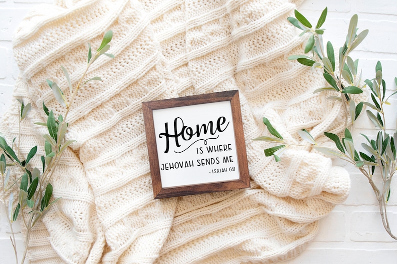 May include: A square wooden sign with the text "Home is where Jehovah sends me - Isaiah 68" in black script. The sign is set against a cream-colored knitted blanket, with sprigs of greenery on either side.