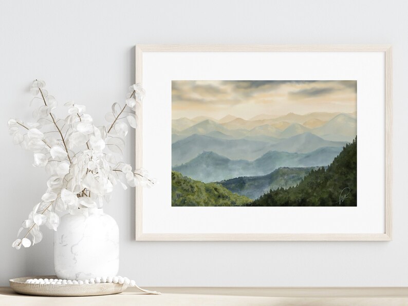 以下が含まれることがあります： 青と緑のぼやけた色合いの山脈を描いた、額縁に入った水彩画。山々は重なり合っており、手前の山はより詳細に描かれ、奥の山は背景に溶け込んでいく。
