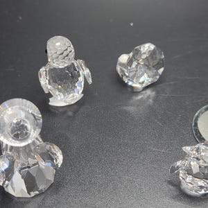 May include: A collection of clear crystal figurines, including a penguin, a heart, and a candle holder. The figurines are faceted, reflecting light. The background is a dark, neutral surface.