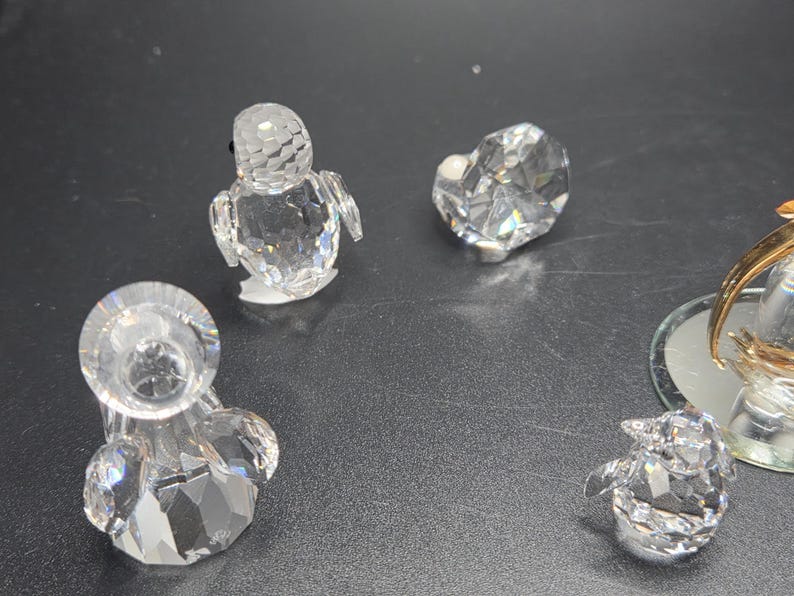 May include: A collection of clear crystal figurines, including a penguin, a heart, and a candle holder. The figurines are faceted, reflecting light. The background is a dark, neutral surface.