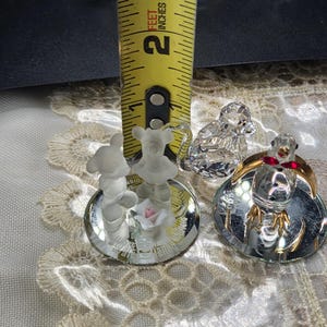 May include: A collection of crystal figurines, including a bear, a monkey, and a bird. The bear figurine is approximately 1 inch tall. The bird figurine has a gold-colored base and red accents. The figurines are displayed on a decorative surface.