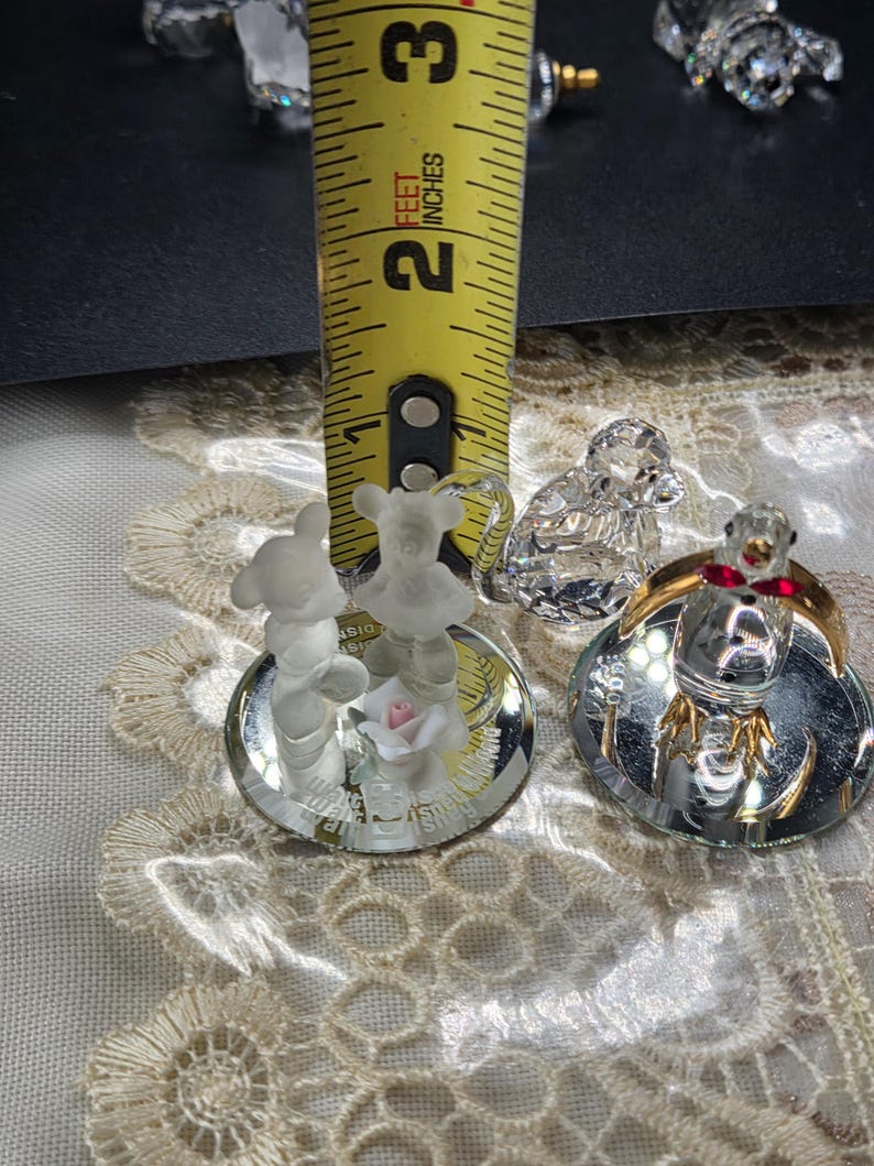 May include: A collection of crystal figurines, including a bear, a monkey, and a bird. The bear figurine is approximately 1 inch tall. The bird figurine has a gold-colored base and red accents. The figurines are displayed on a decorative surface.