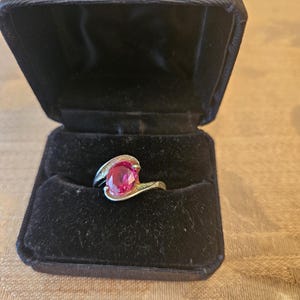 May include: A ring with a pink oval-shaped gemstone set in a gold band. The ring is in a black velvet ring box.