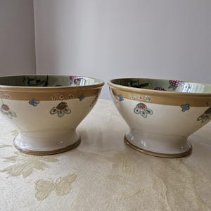 May include: Two white ceramic bowls with a brown rim and floral pattern. The bowls have a pedestal base.
