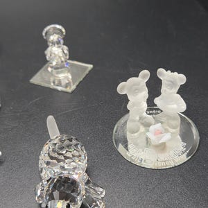 May include: A collection of crystal figurines, including a dog, a chef, and a pair of Mickey and Minnie Mouse figures. The dog is lying down, and the Mickey and Minnie figures are standing on a mirrored base with a small pink rose.
