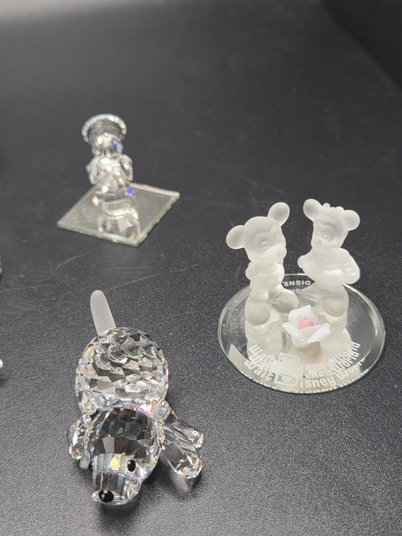 May include: A collection of crystal figurines, including a dog, a chef, and a pair of Mickey and Minnie Mouse figures. The dog is lying down, and the Mickey and Minnie figures are standing on a mirrored base with a small pink rose.