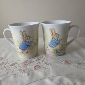 May include: Two white ceramic mugs featuring a classic illustration of a brown rabbit wearing a blue jacket. The rabbit is depicted in a garden setting with soft green and pink floral accents. Each mug has a curved handle and a smooth finish.