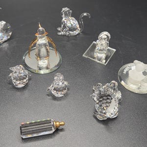 May include: A collection of crystal figurines, including a monkey, a baby, a frog, two penguins, a flower, a bottle, and a perfume bottle with gold accents. The figurines are displayed on a dark surface.