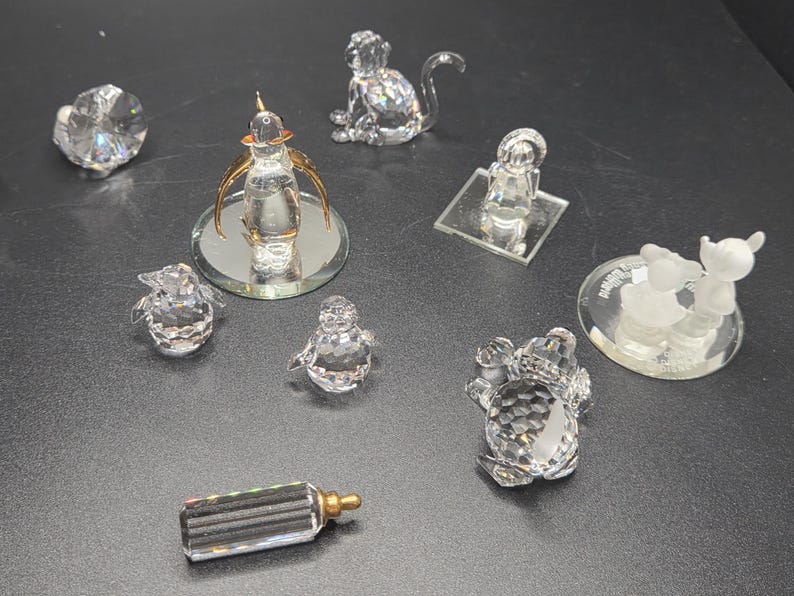 May include: A collection of crystal figurines, including a monkey, a baby, a frog, two penguins, a flower, a bottle, and a perfume bottle with gold accents. The figurines are displayed on a dark surface.