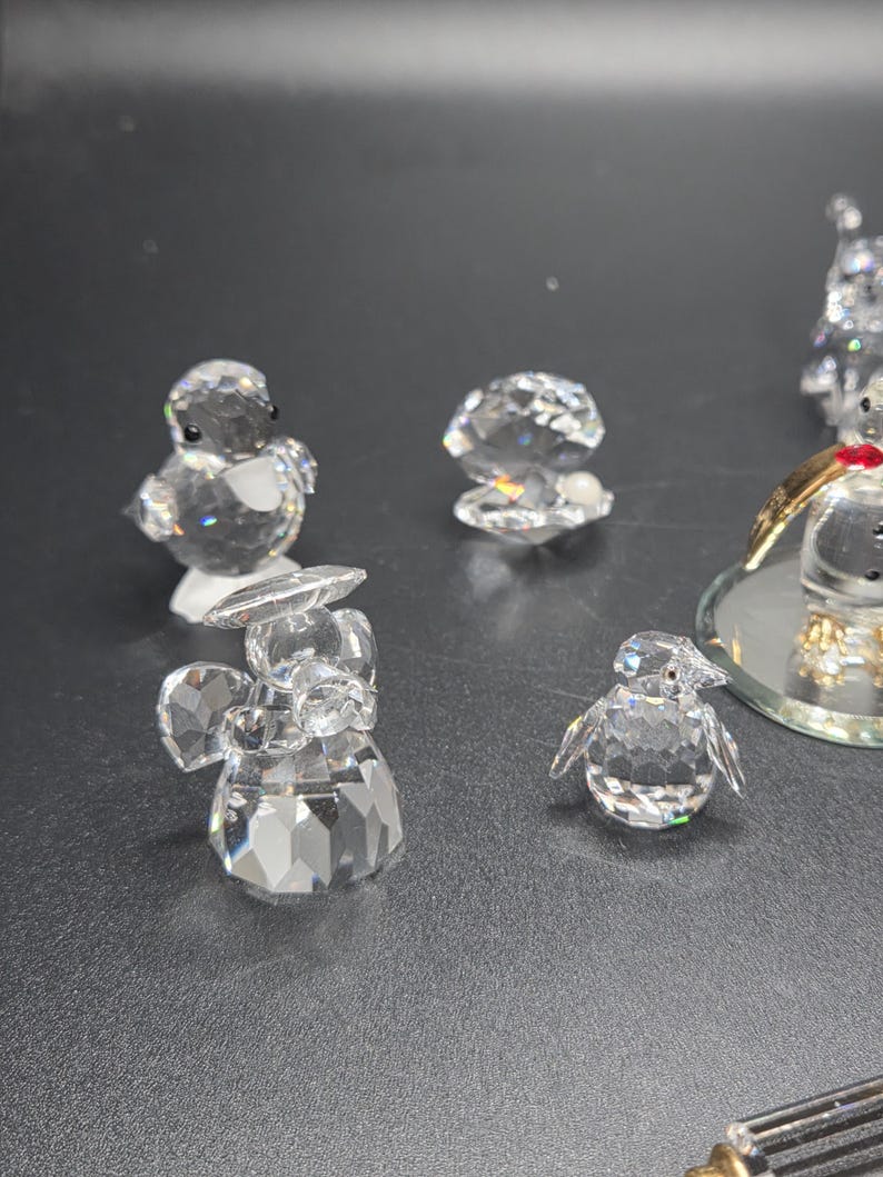 May include: A collection of clear crystal figurines, including a penguin, an angel, and a shell. The figurines are faceted, reflecting light. The penguin has small black eyes. The angel has wings and a halo. The shell is open.