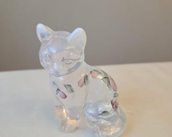 Puede incluir: Una pequeña figura de gato de vidrio translúcido e iridiscente en posición sentada. El gato presenta delicados detalles florales pintados a mano en tonos rosa y verde a lo largo de su espalda y pecho, con un acabado suave y brillante.