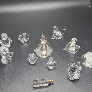 May include: A collection of clear crystal figurines, including a monkey, penguins, a baby bottle, and other decorative objects. The figurines are displayed on a dark surface, showcasing their reflective qualities.