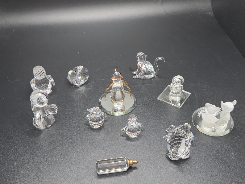 May include: A collection of clear crystal figurines, including a monkey, penguins, a baby bottle, and other decorative objects. The figurines are displayed on a dark surface, showcasing their reflective qualities.