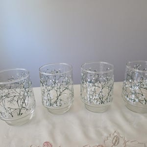 May include: Four clear glass tumblers with a delicate floral design. The glasses feature a pattern of white flowers and green stems, creating an elegant and airy aesthetic. The glasses are arranged on a light-colored surface.