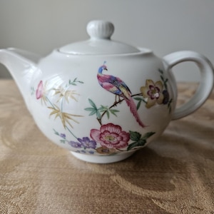May include: White teapot with a floral and bird design. The teapot has a lid and a handle. The flowers are pink, purple, and yellow. The bird is blue, green, and red.