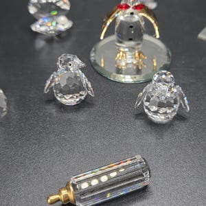 May include: A collection of crystal figurines, including penguins, a baby bottle, and a seashell. The penguins have gold-colored wings and red accents. The baby bottle has a gold-colored nipple. The figurines are displayed on a dark surface.