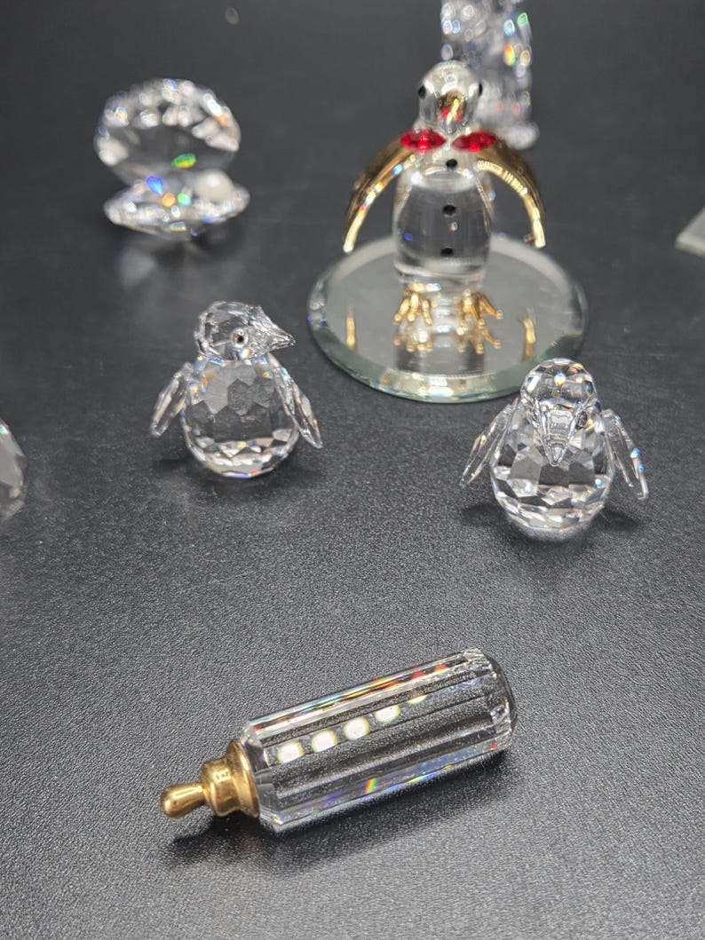 May include: A collection of crystal figurines, including penguins, a baby bottle, and a seashell. The penguins have gold-colored wings and red accents. The baby bottle has a gold-colored nipple. The figurines are displayed on a dark surface.