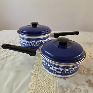 May include: Two blue and white enamel pots with black handles and lids. The pots have a decorative pattern around the base and are sitting on a floral tablecloth. The lids have black knobs.