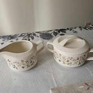 May include: Cream-colored ceramic creamer and lidded sugar bowl set. Both pieces feature a speckled finish and are adorned with a delicate floral pattern in shades of brown, yellow, and green. The creamer has a spout and handle.