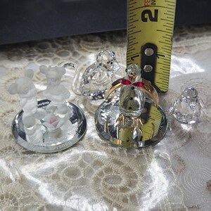 May include: A collection of crystal figurines, including a Mickey and Minnie Mouse pair, a snowman with a red bow, and a small bird. The figurines are displayed on a reflective surface. A yellow measuring tape shows a 2-inch measurement.