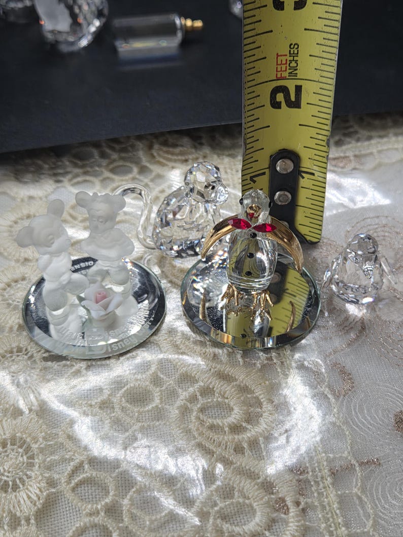 May include: A collection of crystal figurines, including a Mickey and Minnie Mouse pair, a snowman with a red bow, and a small bird. The figurines are displayed on a reflective surface. A yellow measuring tape shows a 2-inch measurement.