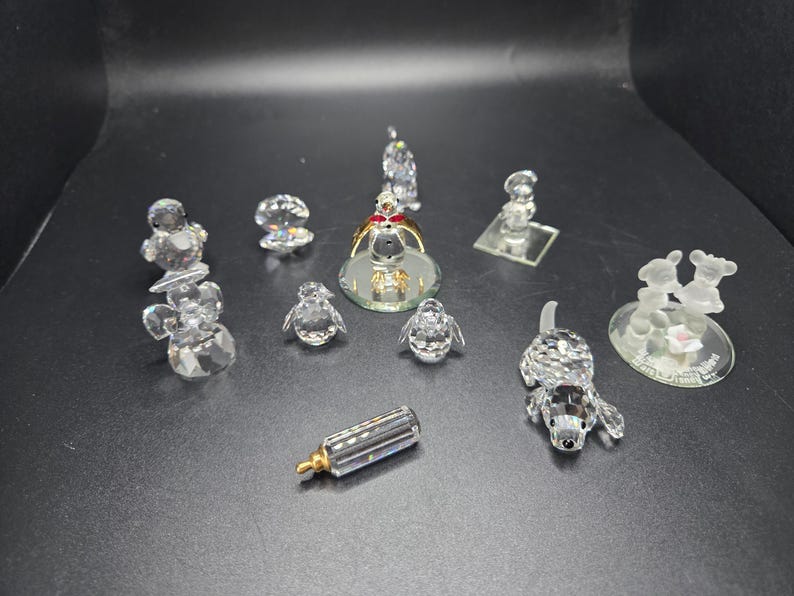 May include: A collection of crystal figurines, including angels, penguins, a dog, and a baby bottle. The figurines are clear and faceted, reflecting light. The collection is displayed on a dark surface.