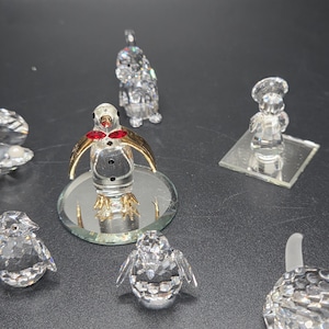 May include: A collection of crystal figurines, including penguins, a chef, and other decorative pieces. The penguin figurines have gold-colored wings and red accents. The figurines are displayed on a dark surface.