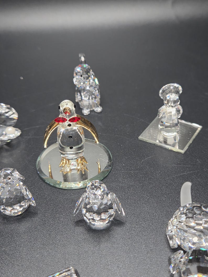 May include: A collection of crystal figurines, including penguins, a chef, and other decorative pieces. The penguin figurines have gold-colored wings and red accents. The figurines are displayed on a dark surface.
