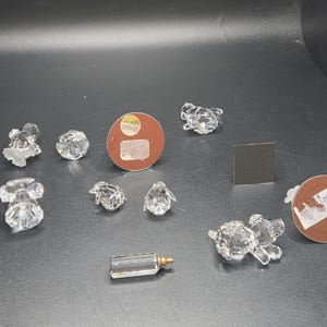 May include: A collection of clear crystal figurines, including a mouse, a frog, a penguin, a baby bottle, and a dog. The figurines are arranged on a dark surface. There are also two round brown stickers and a small square black sticker.