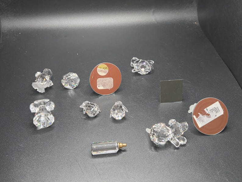 May include: A collection of clear crystal figurines, including a mouse, a frog, a penguin, a baby bottle, and a dog. The figurines are arranged on a dark surface. There are also two round brown stickers and a small square black sticker.