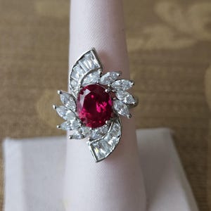 May include: A silver-toned ring featuring a large, oval-cut, ruby-red gemstone at the center, surrounded by smaller, clear, baguette-cut and marquise-cut stones. The ring is displayed on a white ring holder.