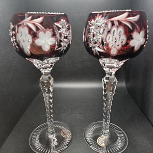 May include: Two ruby red wine glasses with intricate floral and leaf designs. The clear stems have a textured, twisted appearance, and the bases are circular with a ridged pattern. The glasses are likely made of crystal.