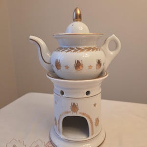 May include: A white ceramic teapot with a gold lid finial, resting on a matching white ceramic warmer base. Both pieces feature gold-colored beetle and star motifs, with gold trim along the rims, handle, spout, and the base's arched opening.