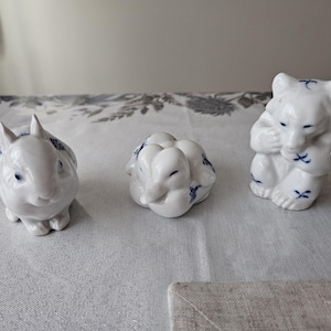 May include: Three white ceramic animal figurines: a rabbit, a duck, and a bear. Each figurine has blue accents and is in a different pose. The rabbit is upright, the duck is curled up, and the bear is sitting.