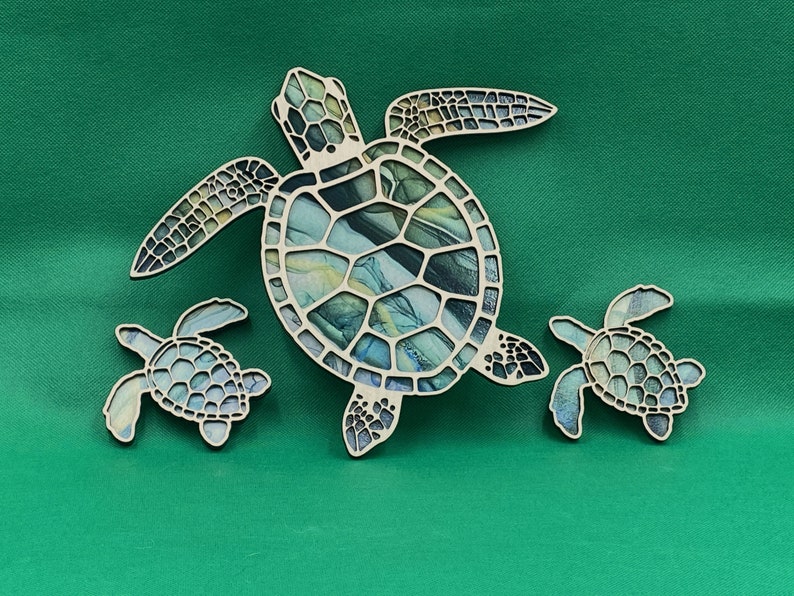 Puede incluir: Tres adornos de madera en forma de tortuga marina, uno grande y dos peque&ntilde;os. La tortuga grande tiene un dise&ntilde;o marmoleado azul, verde y amarillo. Las tortugas m&aacute;s peque&ntilde;as son de un color azul verdoso s&oacute;lido.