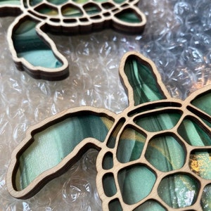 Puede incluir: Dos adornos de tortuga de madera con un dise&ntilde;o de m&aacute;rmol verde y dorado. Las tortugas est&aacute;n recortadas y tienen un patr&oacute;n de caparaz&oacute;n detallado.