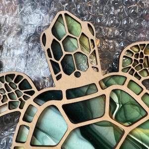 Puede incluir: Un recorte de tortuga de madera con un dise&ntilde;o abstracto en verde, azul y dorado. El caparaz&oacute;n de la tortuga est&aacute; intrincadamente detallado con un patr&oacute;n geom&eacute;trico.