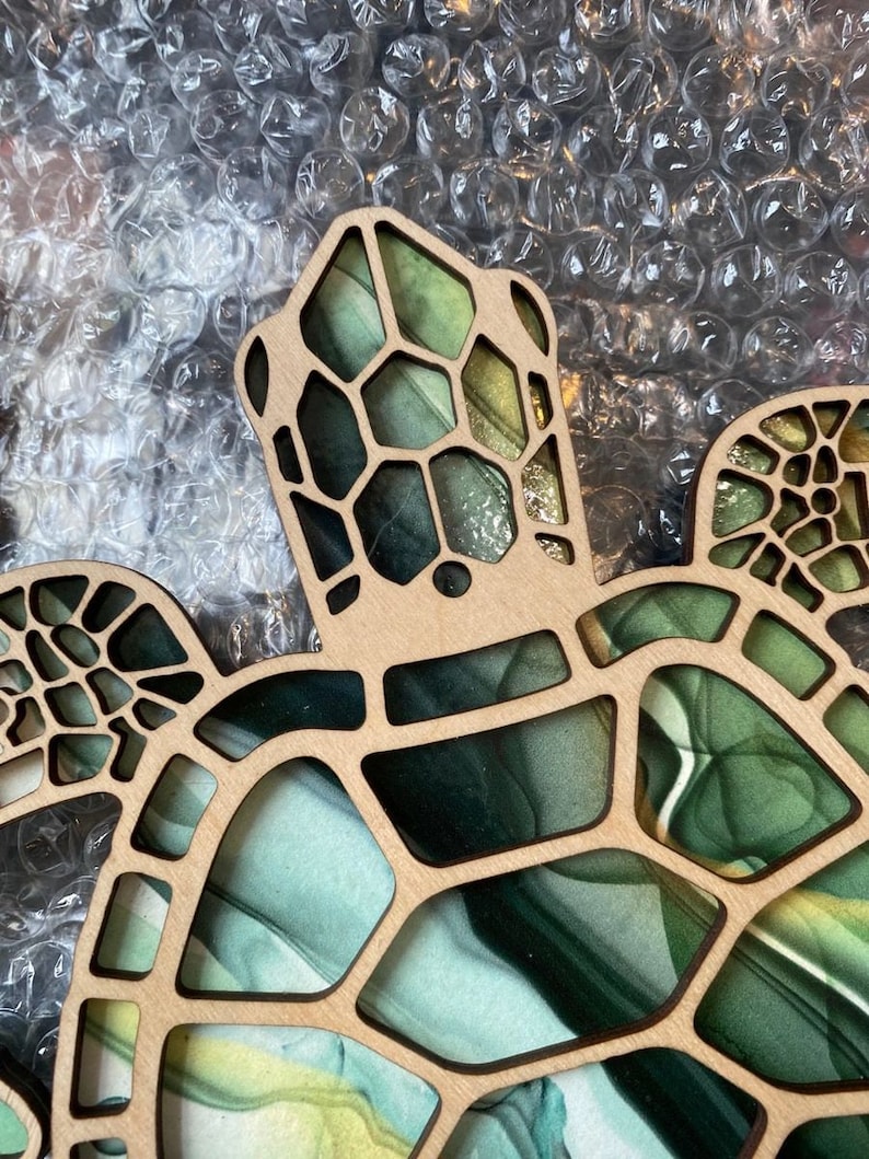 Puede incluir: Un recorte de tortuga de madera con un dise&ntilde;o abstracto en verde, azul y dorado. El caparaz&oacute;n de la tortuga est&aacute; intrincadamente detallado con un patr&oacute;n geom&eacute;trico.