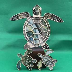 Puede incluir: Un recorte de madera de tres tortugas marinas. La tortuga m&aacute;s grande est&aacute; en el centro y tiene un caparaz&oacute;n con estampado verde, azul y amarillo. Las dos tortugas m&aacute;s peque&ntilde;as est&aacute;n a cada lado de la tortuga grande y tienen un caparaz&oacute;n con un estampado similar.