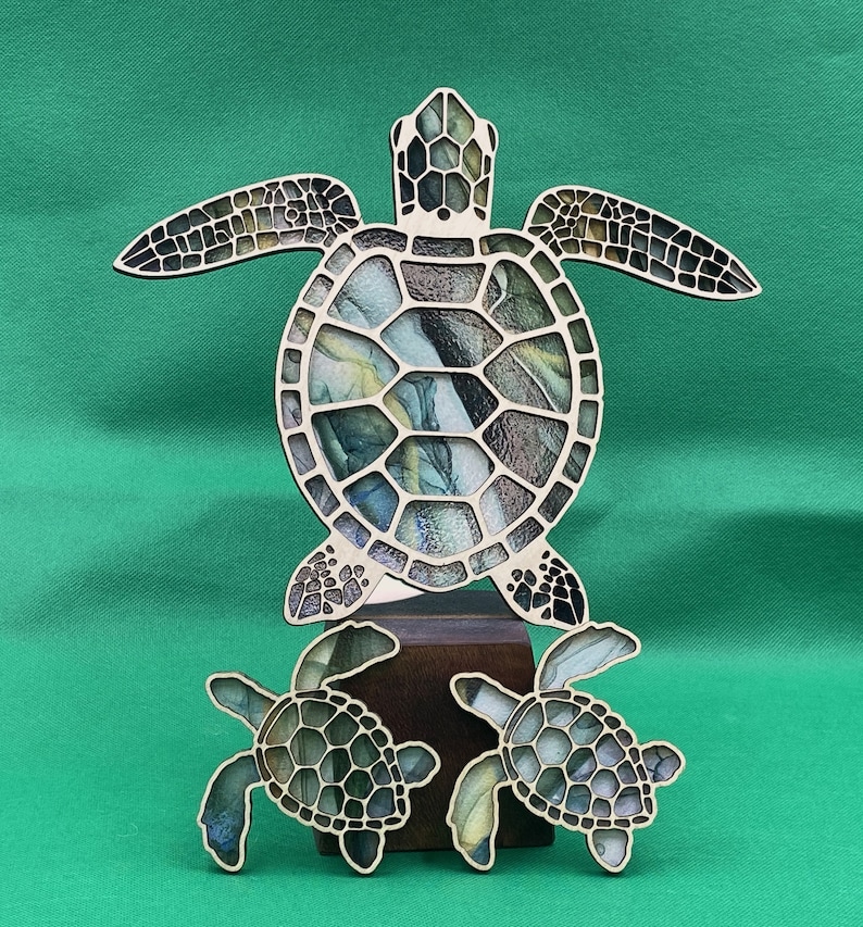 Puede incluir: Un recorte de madera de tres tortugas marinas. La tortuga m&aacute;s grande est&aacute; en el centro y tiene un caparaz&oacute;n con estampado verde, azul y amarillo. Las dos tortugas m&aacute;s peque&ntilde;as est&aacute;n a cada lado de la tortuga grande y tienen un caparaz&oacute;n con un estampado similar.